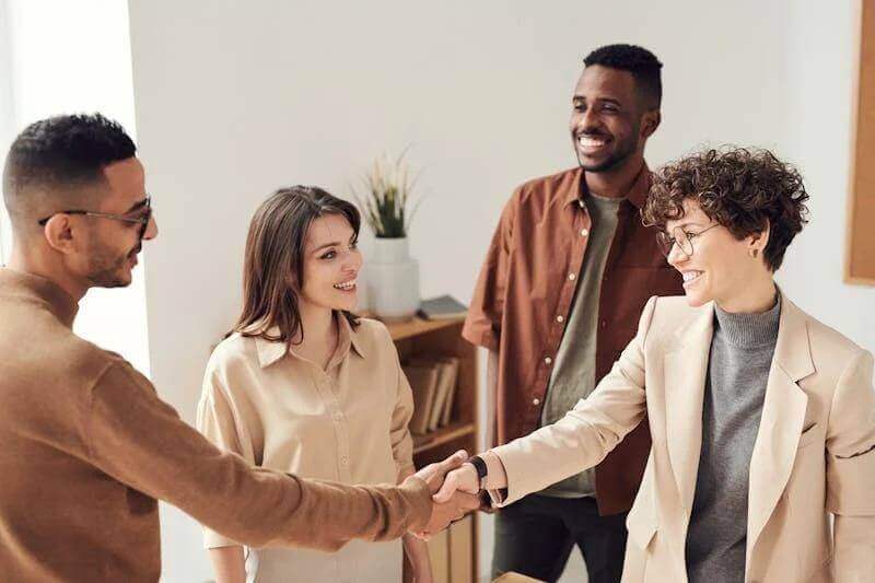 Four people are standing together in an office, smiling. Two of them, a man and a woman, are shaking hands while the other two look on, appearing friendly and engaged. A plant is visible in the background.