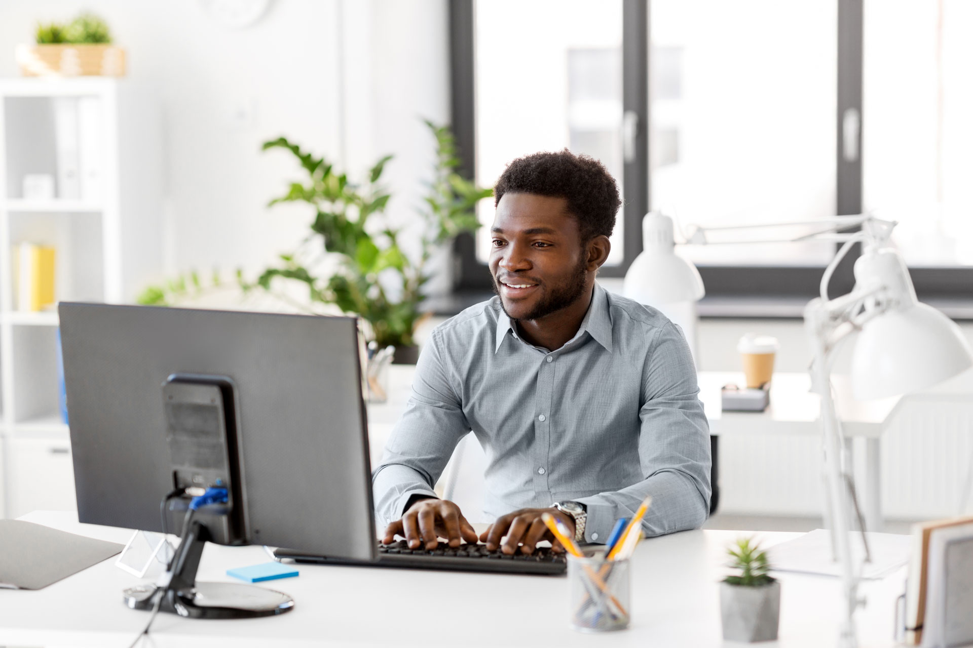 A man in a gray shirt smiles while typing on a keyboard at an office desk with a monitor, desk lamp, and various office supplies. Bright, modern workspace with plants and large windows in the background.
