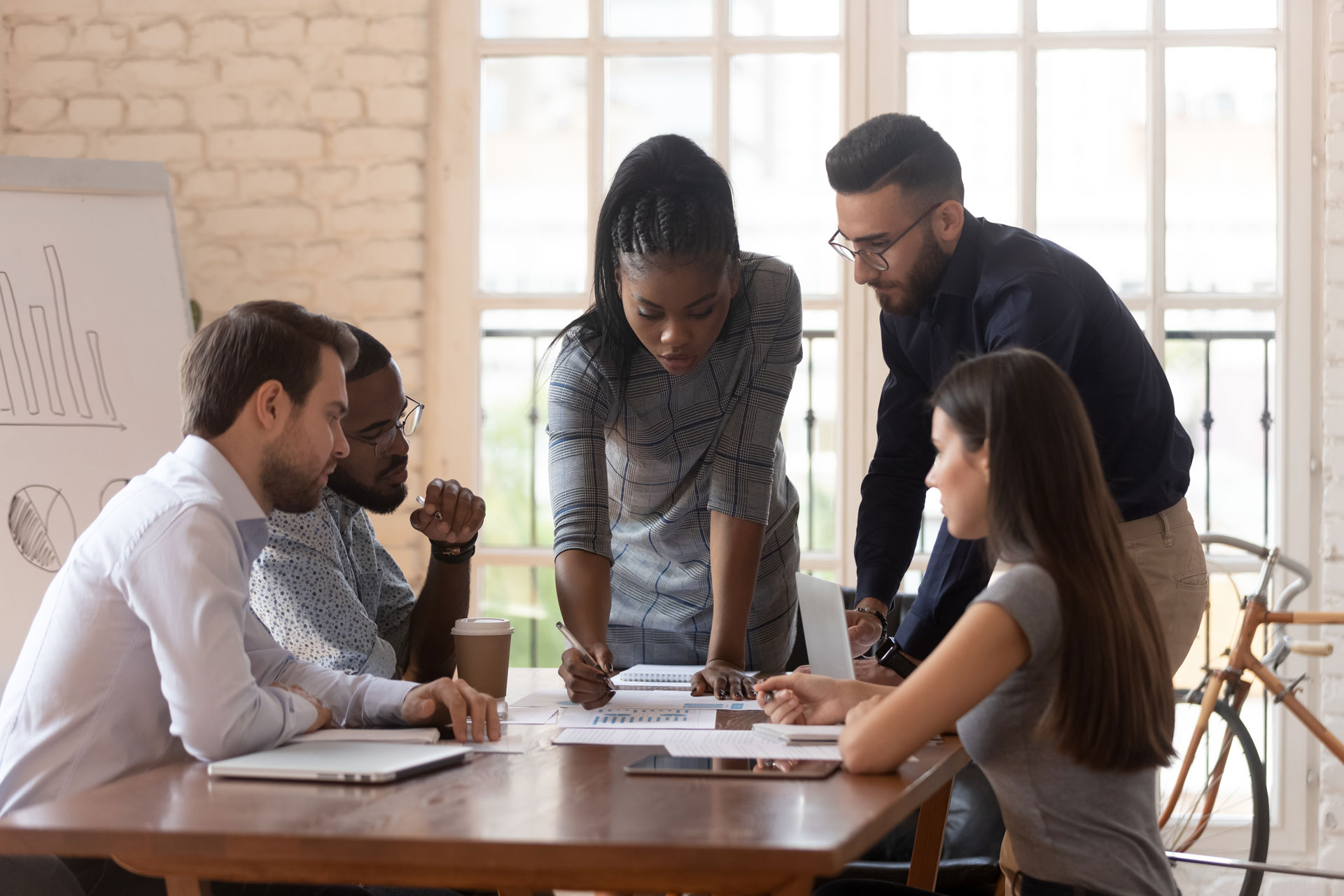 Five people sit and stand around a table, collaborating and reviewing documents. A woman in the center is pointing to a paper while others listen. There is a whiteboard, coffee cup, and laptops in the bright office setting.