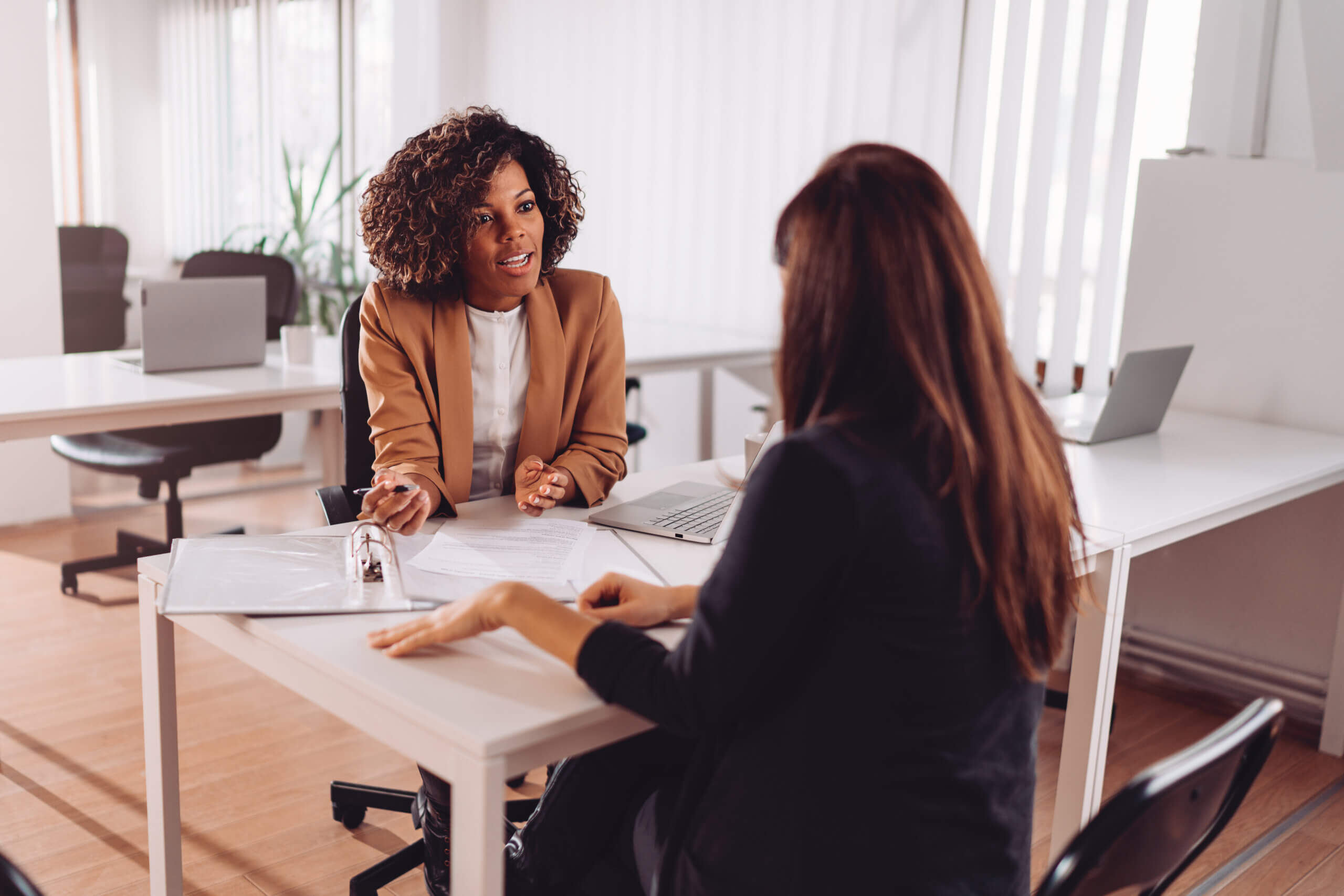 Two women sit across from each other at a desk in a modern office, engaged in a conversation. One woman takes notes while the other gestures as she speaks. Papers, a binder, and a laptop are on the desk.