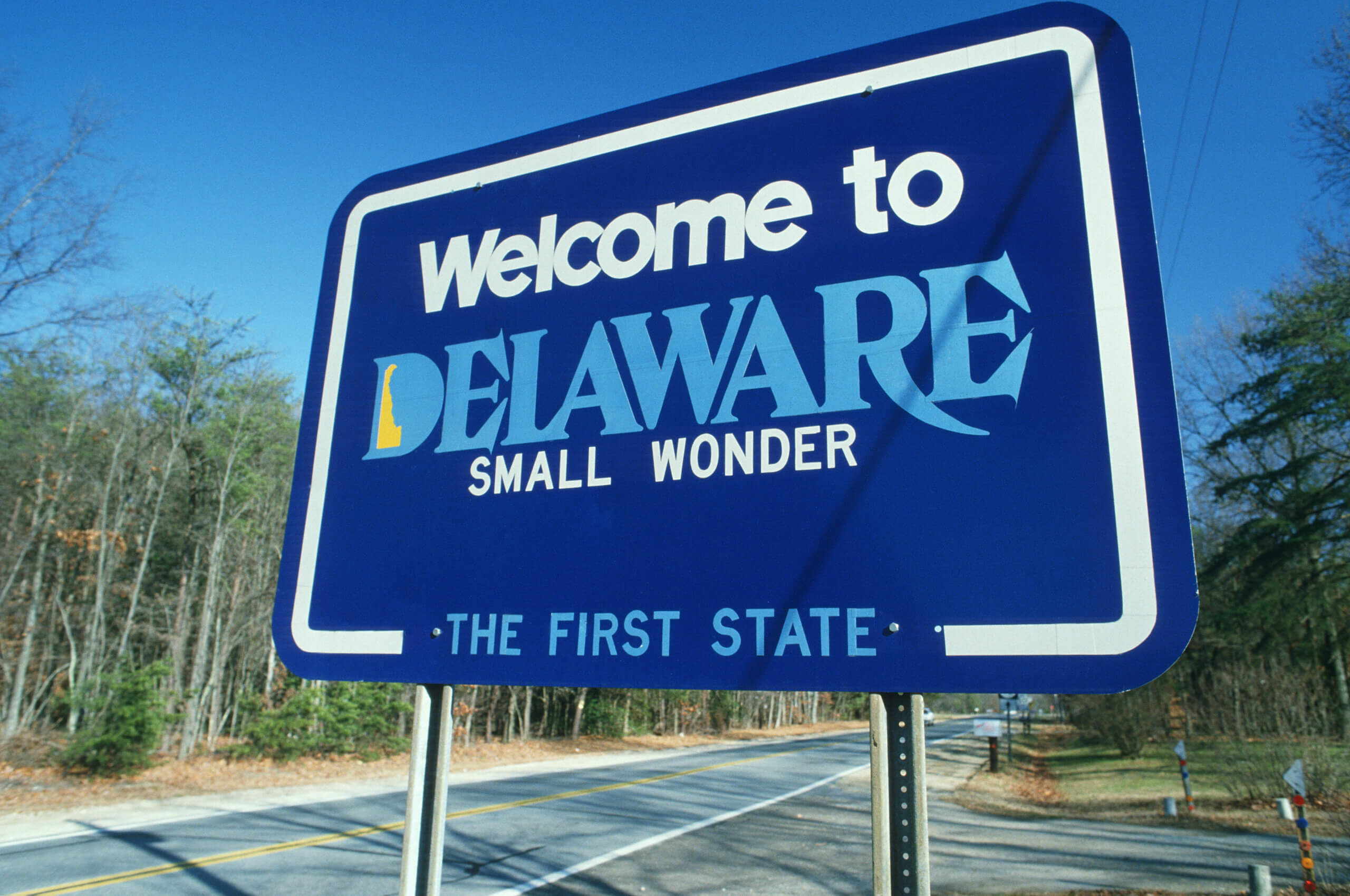 A blue roadside sign reads "Welcome to Delaware, Small Wonder, The First State," with trees and a road visible in the background.