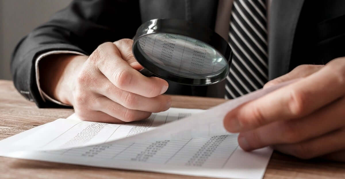 A person in a suit examines financial documents on a desk with a magnifying glass, closely inspecting rows of numbers on the papers.