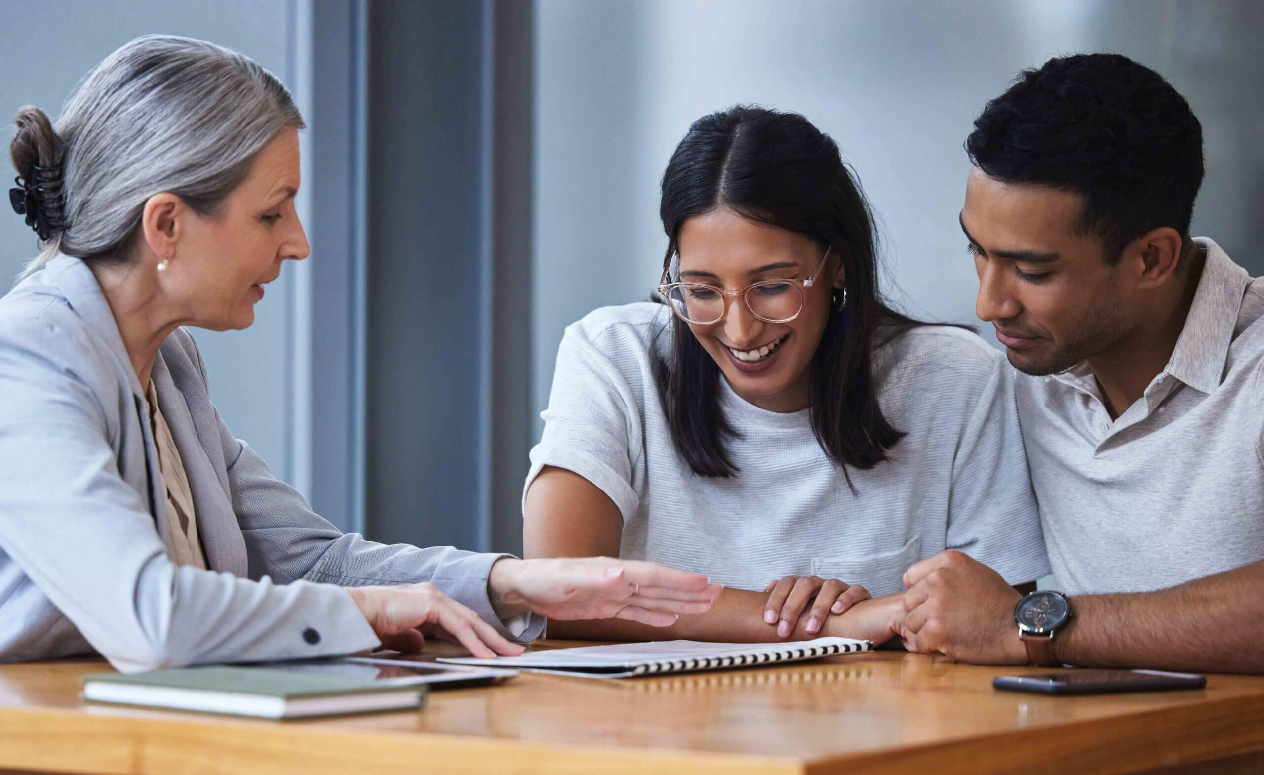 A woman in a blazer sits at a table with a smiling young couple, showing them a document. The couple, both in casual clothes, look engaged and interested as they review the papers together.