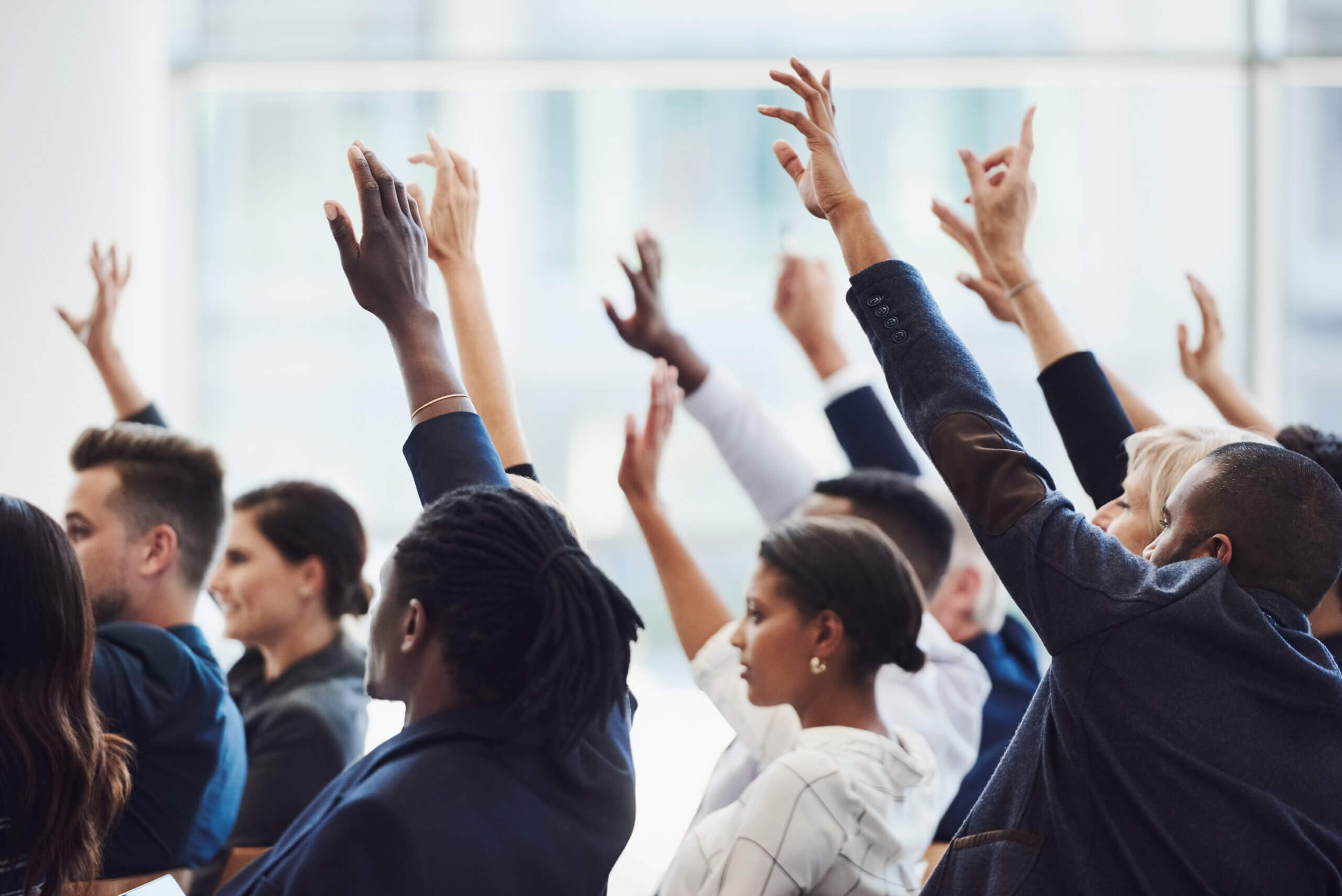 A diverse group of people sits in rows, raising their hands enthusiastically, as if participating or volunteering during a meeting or conference in a bright, modern room.