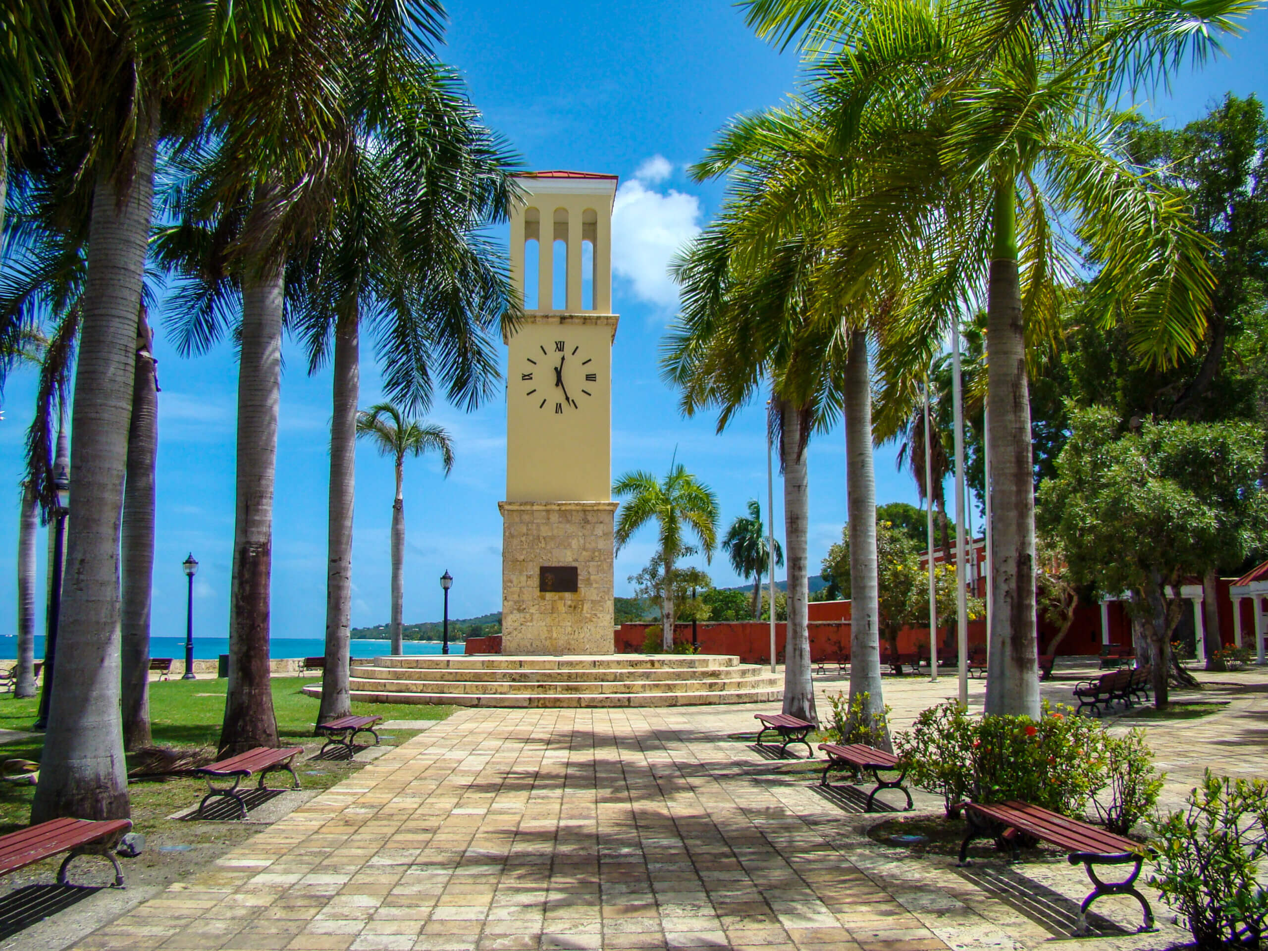 A tall clock tower stands in a sunny plaza surrounded by palm trees, benches, and lush greenery, with a bright blue sky and the ocean visible in the background.