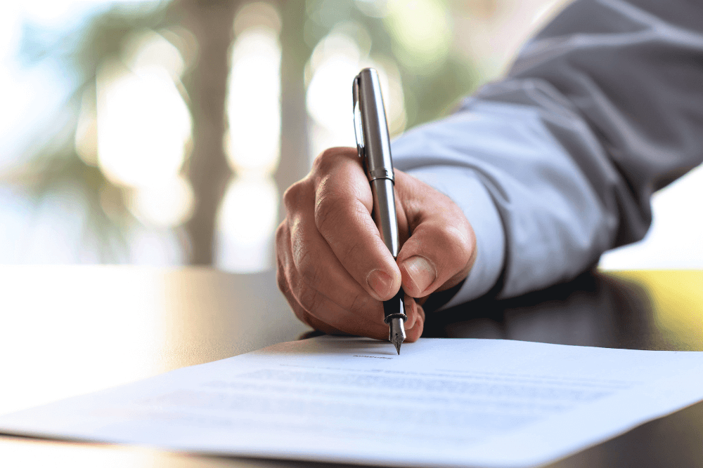 A person in a long-sleeve shirt signing a document with a silver pen on a dark table, with a blurred background.