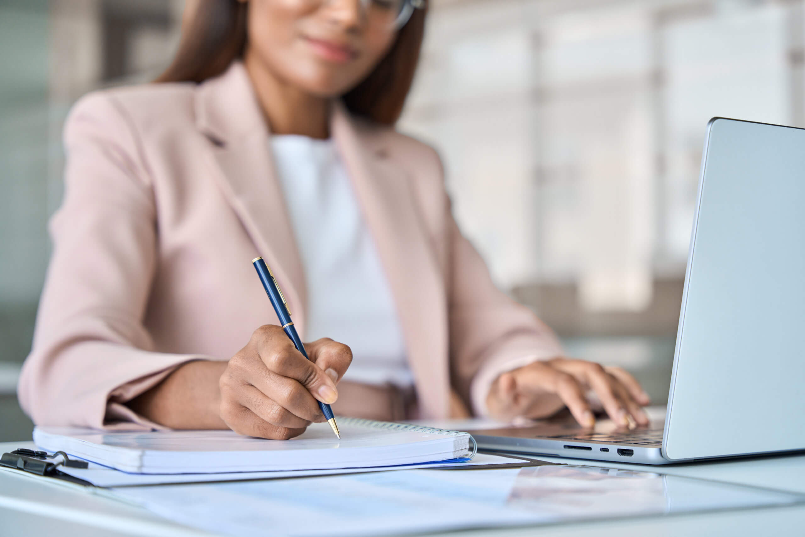 A woman in a beige blazer writes in a notebook with a pen while using a laptop. A clipboard and pen rest on the desk. The setting appears to be a bright, modern office. Her face is partially out of focus.