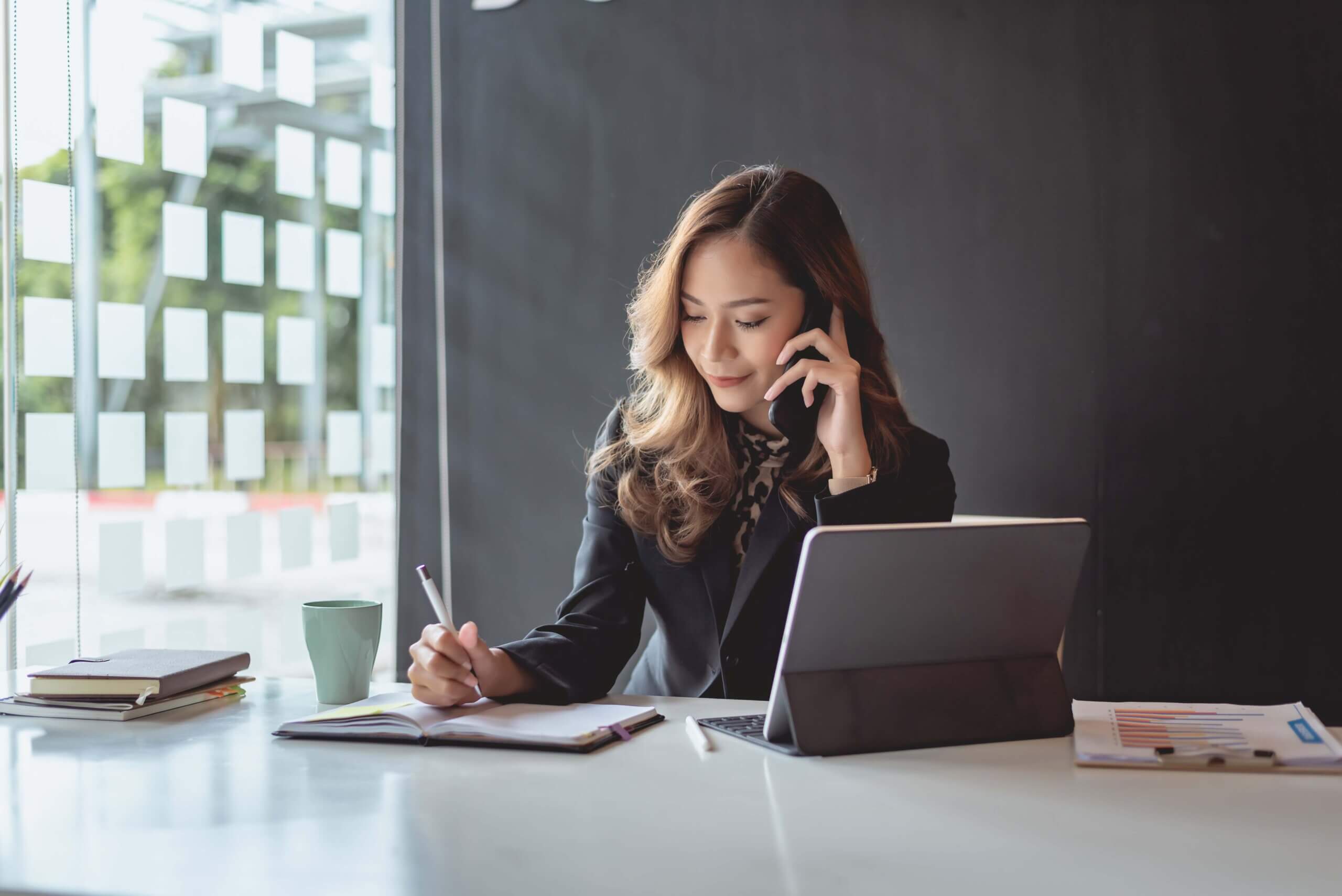 A woman sits at a desk with a laptop, talking on the phone and writing in a notebook. She wears business attire and is working near a large window with natural light. A coffee mug and documents are on the desk.