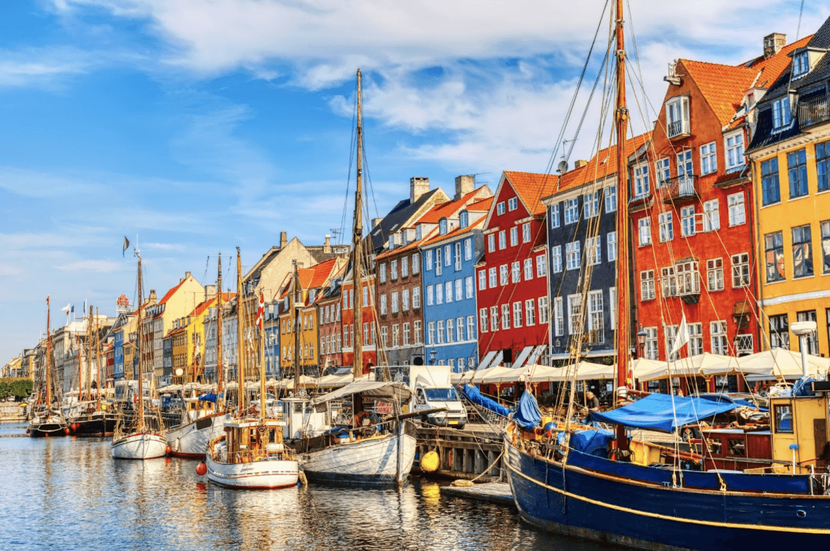 Colorful row houses line a waterfront street in Nyhavn, Copenhagen, Denmark. Wooden boats are docked along the canal on a sunny day with blue sky and scattered clouds.