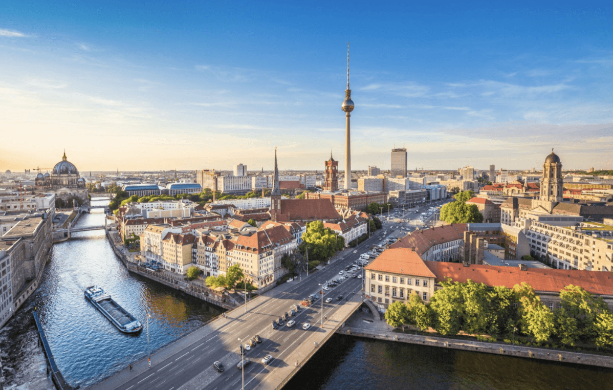 Aerial view of central Berlin, Germany, featuring the River Spree, a large TV tower, historic buildings, bridges, and city streets under a clear blue sky.