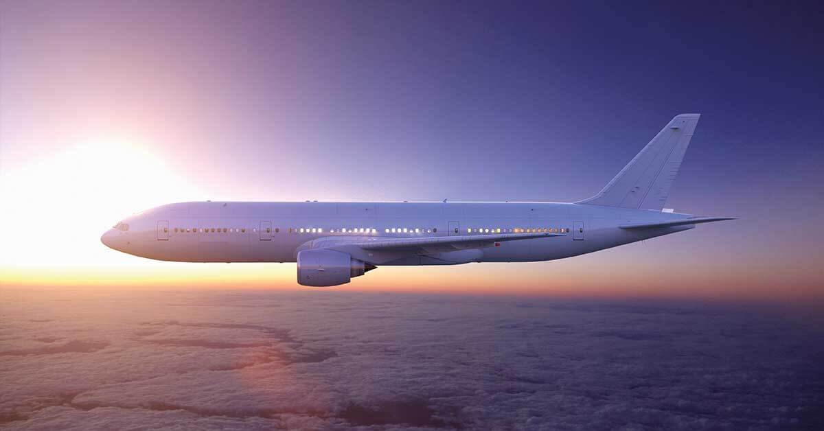 A commercial airplane flying above the clouds at sunset, with the sun glowing on the horizon and the sky tinted with shades of purple and orange.