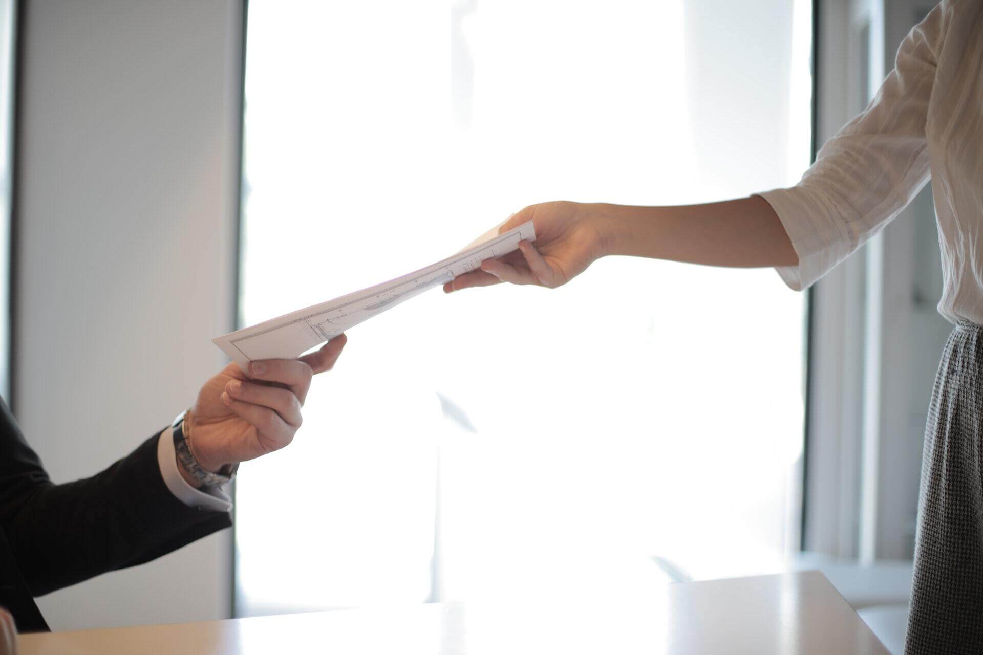 A person standing hands a document to another seated person across a desk in a bright office setting. Only their arms are visible.