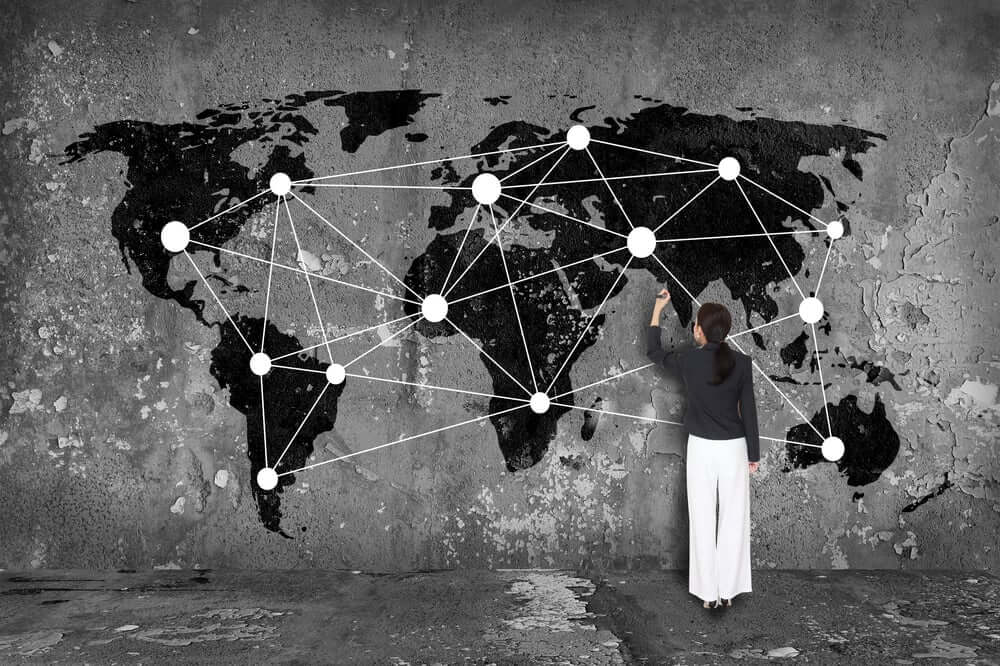 A woman stands in front of a large world map painted on a concrete wall, drawing white interconnected lines and dots that span across continents, symbolizing a global network.