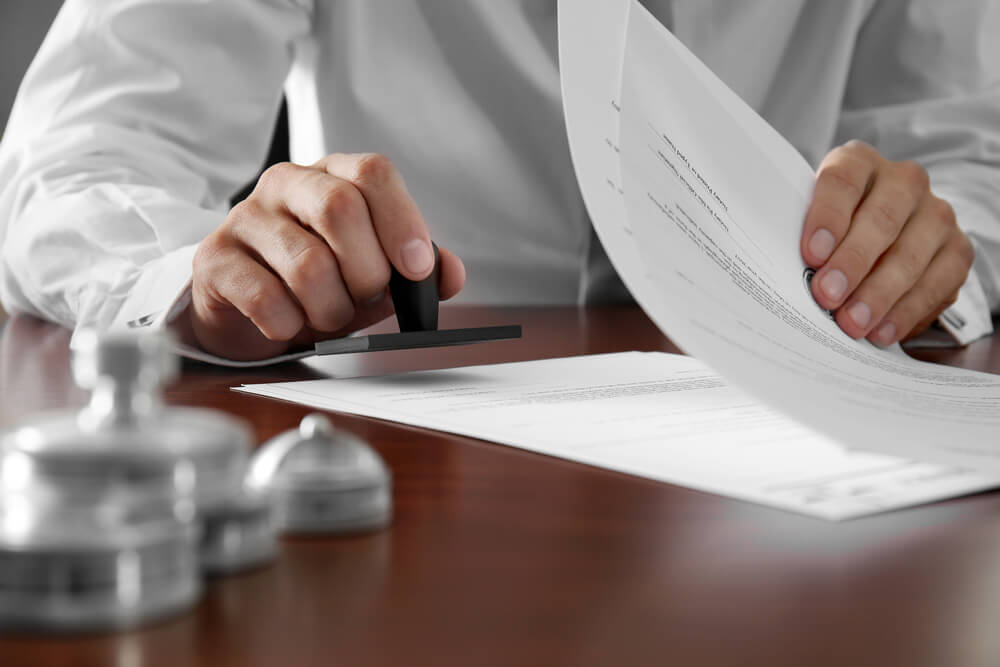 A person in a white shirt holds a rubber stamp and presses it onto a document while holding papers on a wooden desk with other stamps nearby.
