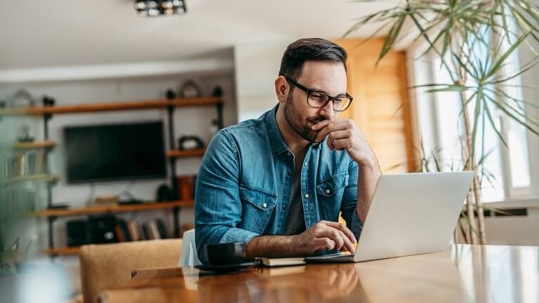 A man wearing glasses and a denim shirt sits at a wooden table, looking thoughtfully at his laptop. A coffee mug is beside him, and the room is bright with natural light and houseplants.