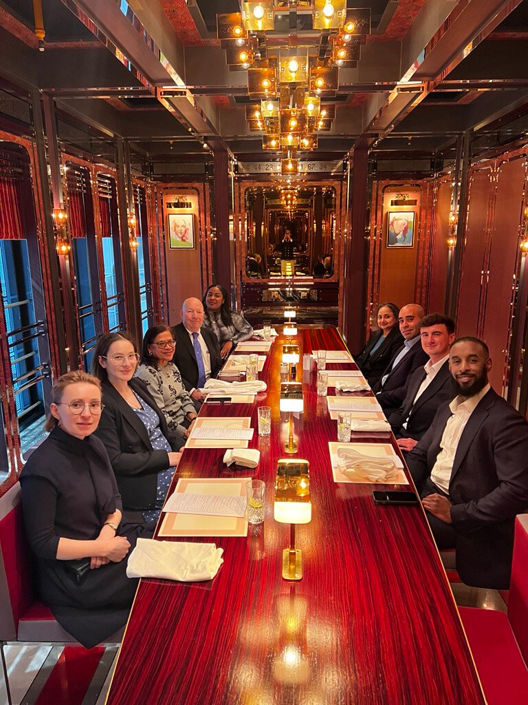 A group of ten people dressed in business attire sit around a long, glossy red table in an elegant, warmly lit restaurant. Menus, napkins, and candles are arranged neatly on the table.