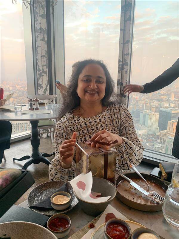 A woman with long dark hair smiles while holding a gift box at a restaurant table with food. Large windows behind her reveal a cityscape under a partly cloudy sky.