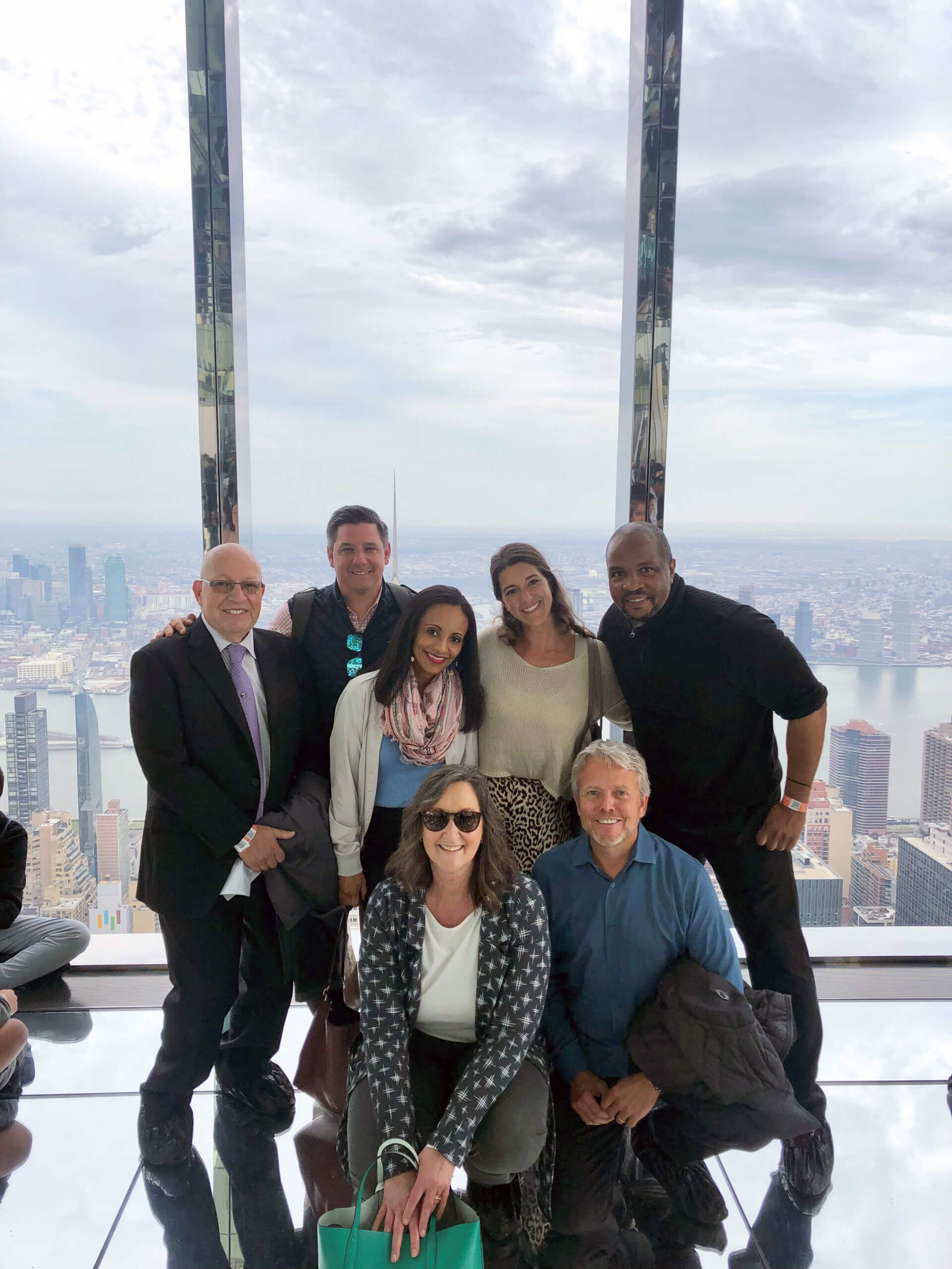 Seven people pose and smile together in front of large windows overlooking a city skyline from a high vantage point on a cloudy day. Some are standing, others are kneeling, and tall buildings are visible outside.