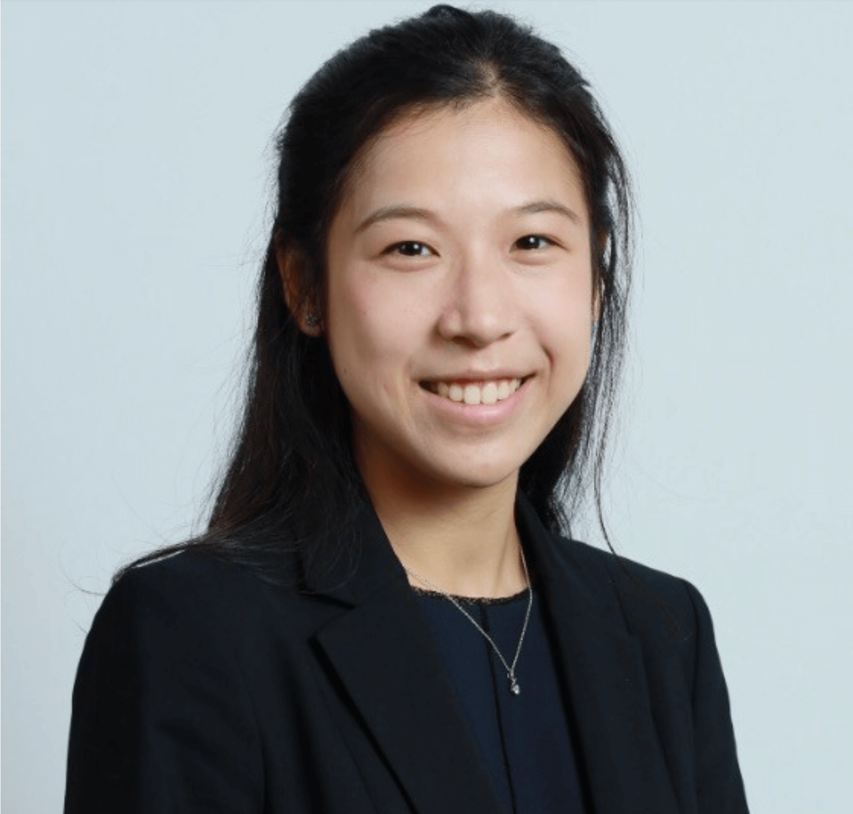 A young woman with long dark hair smiles at the camera. She is wearing a black blazer over a dark top and a delicate necklace, posed against a light grey background.