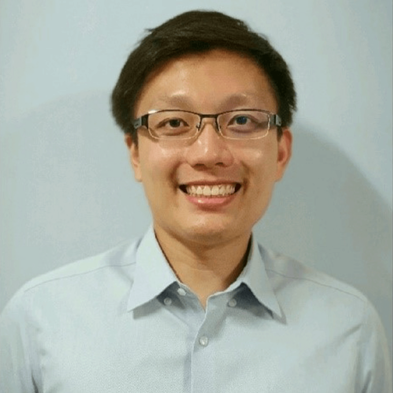 A man with short dark hair and glasses smiles at the camera. He is wearing a light blue button-up shirt and stands in front of a plain light-colored background.