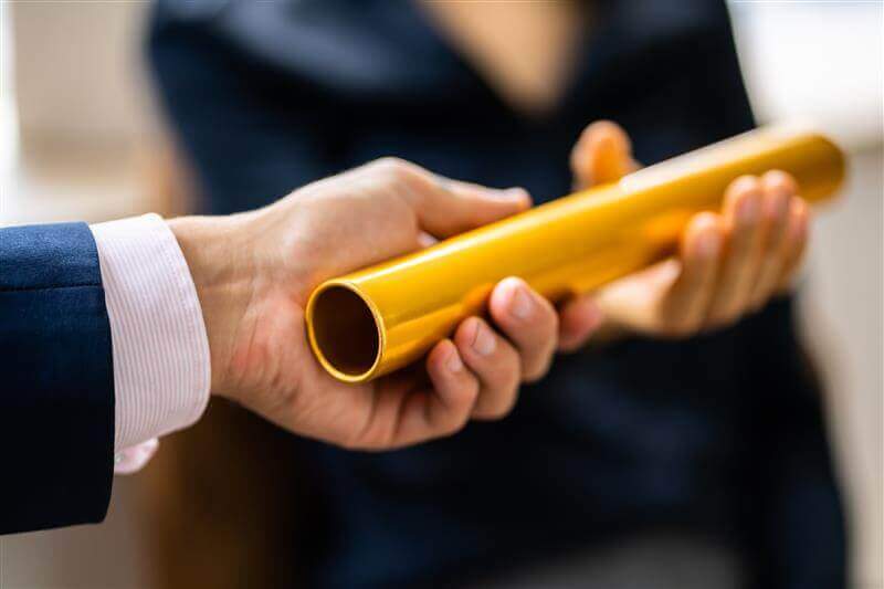 Close-up of one person handing a gold baton to another, symbolizing the concept of passing responsibility or teamwork. Both individuals are wearing business attire.